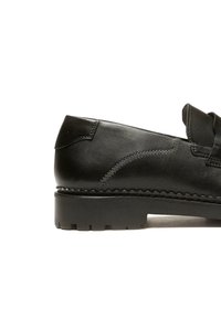 Scarpa in pelle nera con superficie liscia, cuciture sottili e un fiocco decorativo sulla parte superiore. Presenta una suola in gomma robusta e strutturata.