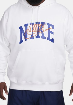 Valkoinen huppari, jossa suuri sininen NIKE-teksti ja päällekkäinen oranssi Nike-swoosh-logo, päällä tumman ihonvärinen henkilö ja tummansiniset housut.