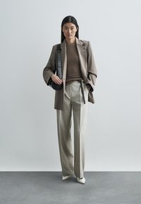 Εξατομικευμένο καφέ σακάκι πάνω από ένα καφέ τοπ, συνδυασμένο με ανοιχτό γκρι παντελόνια φαρδιάς γραμμής. Η εμφάνιση συμπληρώνεται από λευκά μυτερά παπούτσια και μια μικρή μαύρη τσάντα.