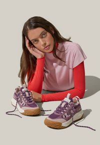Scarpe da ginnastica viola e bianche con tomaia texturizzata, lacci a contrasto e suola in gomma. Il modello posa in modo informale, indossando un top rosa e rosso.