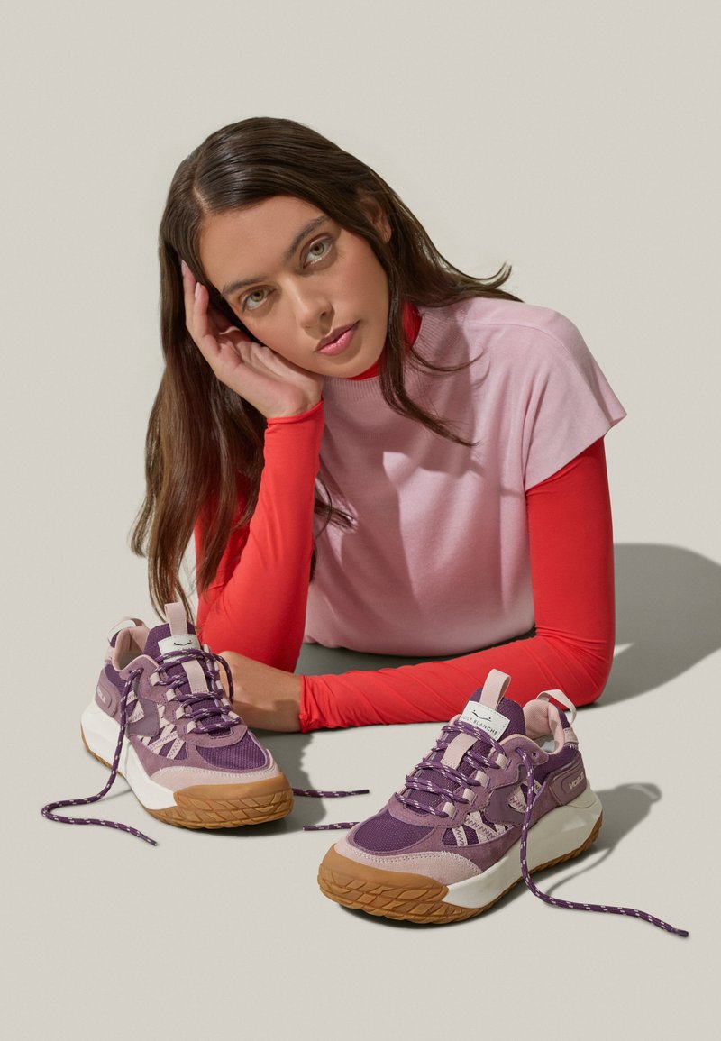 Scarpe da ginnastica viola e bianche con tomaia texturizzata, lacci a contrasto e suola in gomma. Il modello posa in modo informale, indossando un top rosa e rosso.