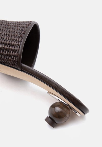 Καφέ υφαντό δερμάτινο mules με μοναδικό στρογγυλό καφέ τακούνι και μπεζ εσωτερική σόλα, φαίνεται από πλάγια γωνία σε λευκό φόντο.