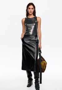 Czarny top z cekinami na ramiączkach z okrągłym dekoltem, w połączeniu z czarną, skórzaną spódnicą o wysokim stanie. Modelka trzyma brązową torbę z kolorowym akcentem.
