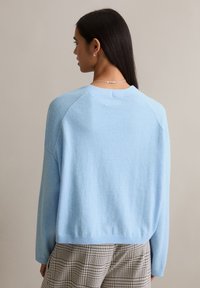 Jasnoniebieski sweter, miękki dzianinowy z okrągłym dekoltem i prążkowanym wykończeniem na dole. Posiada rękawy raglanowe i bezszwowy tył. Widoczne poniżej kraciaste spodnie.