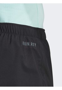 Svarta sportshorts med elastisk midja, med en slät textur och präglad "RAIN.RDY"-logotyp. Ovan syns en ljus mintfärgad tröja.
