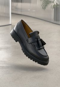 Schwarze Lederloafer mit Quasten-Detail, ausgestattet mit einer strukturierten Sohle und abgerundetem Zehenbereich, vor einem neutralen Hintergrund schwebend.