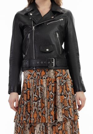 Veste de motard en cuir noir avec plusieurs fermetures éclair argentées et ceinture, portée sur une jupe plissée à plusieurs couches avec un imprimé serpent orange et marron.