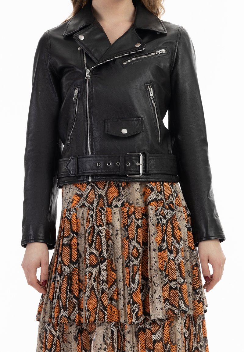 Veste de motard en cuir noir avec plusieurs fermetures éclair argentées et ceinture, portée sur une jupe plissée à plusieurs couches avec un imprimé serpent orange et marron.