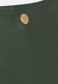 Zaļš maisiņš, izgatavots no teksturēta materiāla, ar apaļu zelta pogu, kurai ir reljefā apdruka, kā aizvēršanas detaļu.