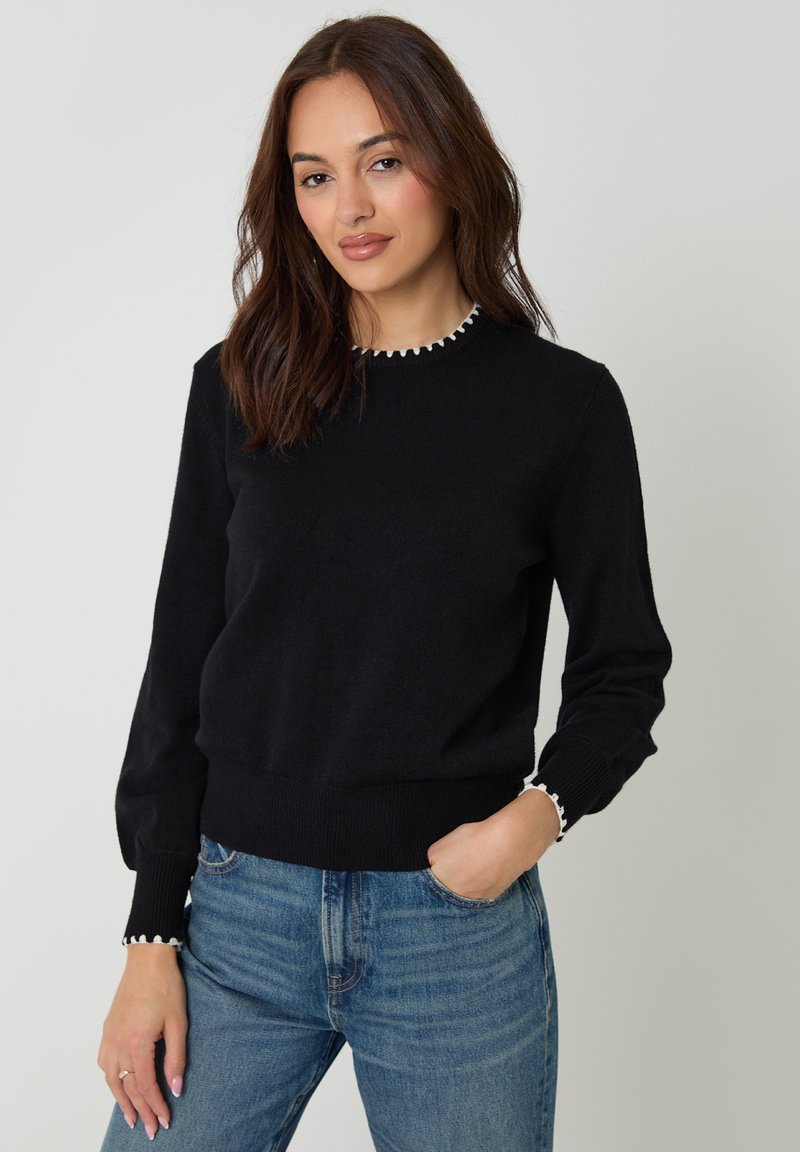 Žena s hnědými vlasy po ramena, oblečená v černém svetru s bílým lemem a modrých džínách, stojící s jednou rukou v kapse.