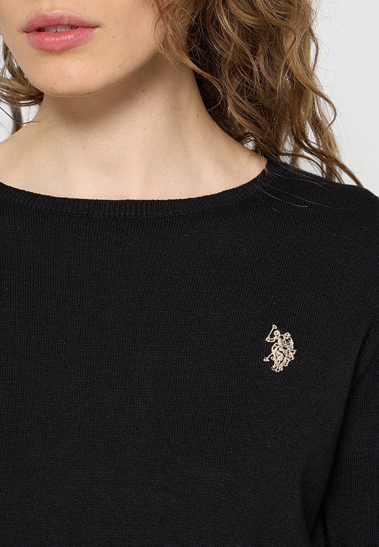 Schwarzer Strickpullover mit rundem Halsausschnitt und kleinem, gesticktem goldenen Emblem auf der linken Brust. Glatte Textur und figurbetontes Design.
