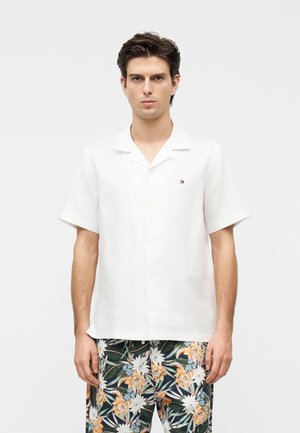 Man draagt een wit overhemd met korte mouwen en bloemenprint broek, staand tegen een effen grijze achtergrond.