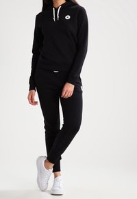 Sweat-shirt à capuche noir avec une poche avant et des cordons blancs, associé à un jogger noir. Porté avec des baskets blanches.