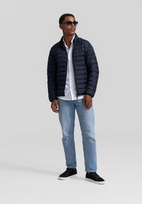 Marineblauwe pufferjack met een rits aan de voorkant, wit shirt, lichtblauwe jeans en zwarte sneakers. Het model draagt donkere zonnebril.