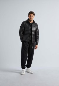 Zwarte gewatteerde jas met een gequilte ontwerp, gecombineerd met zwarte joggingbroeken met "JACKIT" in goud. Het model draagt witte sneakers.