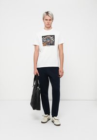 White cotton t-shirt with a graphic print, navy pants, and a black tote bag. Model wears cream sneakers. Simple, modern design.