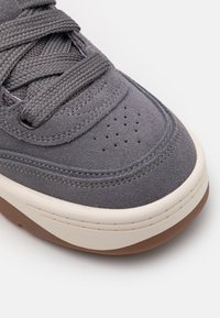 Baskets en daim gris avec surface texturée, bout rond, semelle intermédiaire blanche contrastante et semelle extérieure en caoutchouc marron. Comprend des lacets tissés gris et des perforations.