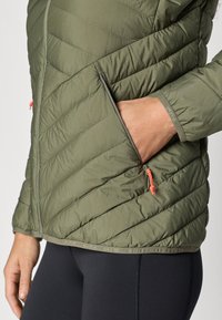 Olīvu zaļa aizšūta jaka ar vertikālām šuvēm, ar rāvējslēdzēja kabatu un oranžiem akcentiem pie rāvējslēdzēja un apakšējā apmale. Nēsāta virs melniem legingiem.