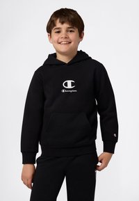 Glunderend kind in een zwart Champion hoodie en bijpassende zwarte sweatpants, staande met ontspannen handen tegen een lichte effen achtergrond.