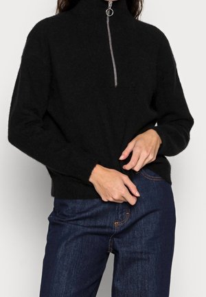 Sort fuzzy pullover med en sølv lynlås ved kraven, båret med mørkeblå jeans, hænder justerer sweaterens kant.