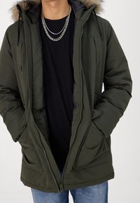 Olivgrön vadderad jacka med pälsfodrad huva, dragkedja, flera fickor framtill och svart innerfoder. Bärs över en svart skjorta.