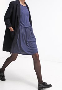 Marint prickig klänning med elastisk midja, tillsammans med en svart lång kappa. Modellen bär svarta skor och genomskinliga svarta strumpbyxor.