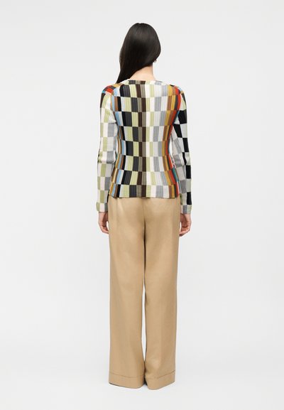 Žena s dlouhými tmavými vlasy, která má na sobě barevný kostkovaný top s dlouhým rukávem a béžové kalhoty s širokými nohavicemi, stojí zády k nám na bílém pozadí.