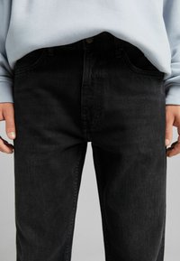 Mustat farkut suoraa leikkausta, joissa on etutaskut ja napitus, käytetty vaalean sinisen neuleen alla.