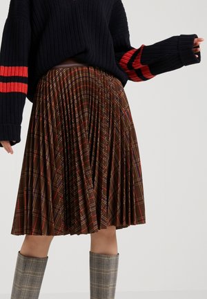 Vrouw draagt een bruine geplooide geruite rok, een marineblauwe trui met rode strepen op de mouwen en grijze geruite kniehoge laarzen tegen een effen achtergrond.