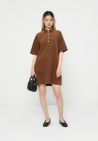 Robe polo en velours côtelé marron avec des manches courtes et des détails de boutons. Associée à un sac à main noir texturé et des chaussures plates noires.