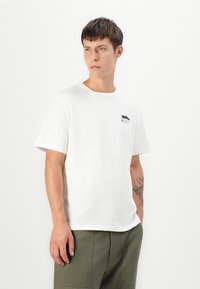 White cotton T-shirt with short sleeves, crew neck, and a small black graphic on the left chest. Worn with olive green pants.