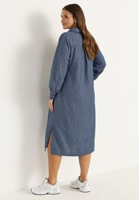 Denim-mekko pitkillä hihoilla, kauluksella ja sivuaukkoilla. Saattaa vaaleansinisestä sävystä ja väljistä mitoista, yhdistetty valkoisiin urheilukenkiin.