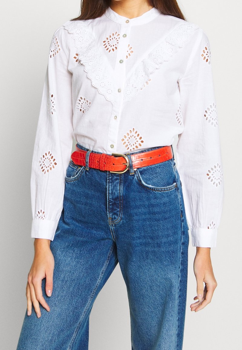 Blouse en coton blanc avec découpes en broderie anglaise, col à volants et boutons perle, associée à un jean en denim bleu taille haute et une ceinture rouge en cuir de crocodile.