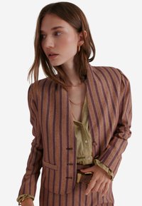 Gestreepte blazer in paars en oranje, gemaakt van gestructureerde stof. Heeft een hoge kraag, drie knopen en een slim fit, gecombineerd met een groene shirt.