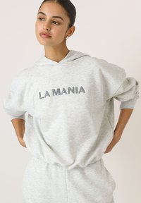 Svetlosivá mikina s kapucňou, na prednej strane so šedým výšivkom "LA MANIA" v tmavšej sivej farbe, vo voľnom strihu a s jemnou textúrou.