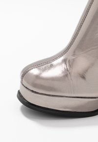 Metalické stříbrné vysoké boty na podpatku s hladkým povrchem, kulatou špičkou a pevným černým platformovým podešví, které zdůrazňují precizní prošívání podél okraje.