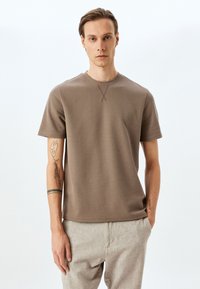 Kortærmet, brun vaffelstrikket T-shirt med rund halsudskæring og diskret tværstingdetalje, kombineret med lysebeige bukser.