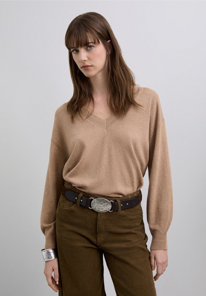 Jersey beige de cuello en V con mangas largas, combinado con pantalones de pana marrón y un cinturón ancho negro con hebilla ovalada plateada.