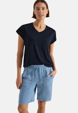 Kvinde iført en sort kortærmet V-hals shirt med subtilt mønster og lyseblå denimshorts med snøre og forlommer.