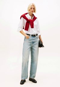 Bela srajca s potiski, svetlo modre kavbojke širokih hlačnic, rdeč pulover, premetan čez ramena, črn pas, črna torbica in natikači z verižico na detajlu.