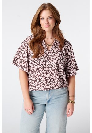 Femme aux longs cheveux roux portant un chemisier floral marron et rose et un jean bleu clair, avec des bijoux en or et une expression neutre.