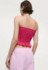 Donna con i capelli corti castani, che indossa un top rosa testurizzato senza spalline e pantaloni larghi rosa chiaro, in piedi contro uno sfondo chiaro e semplice.