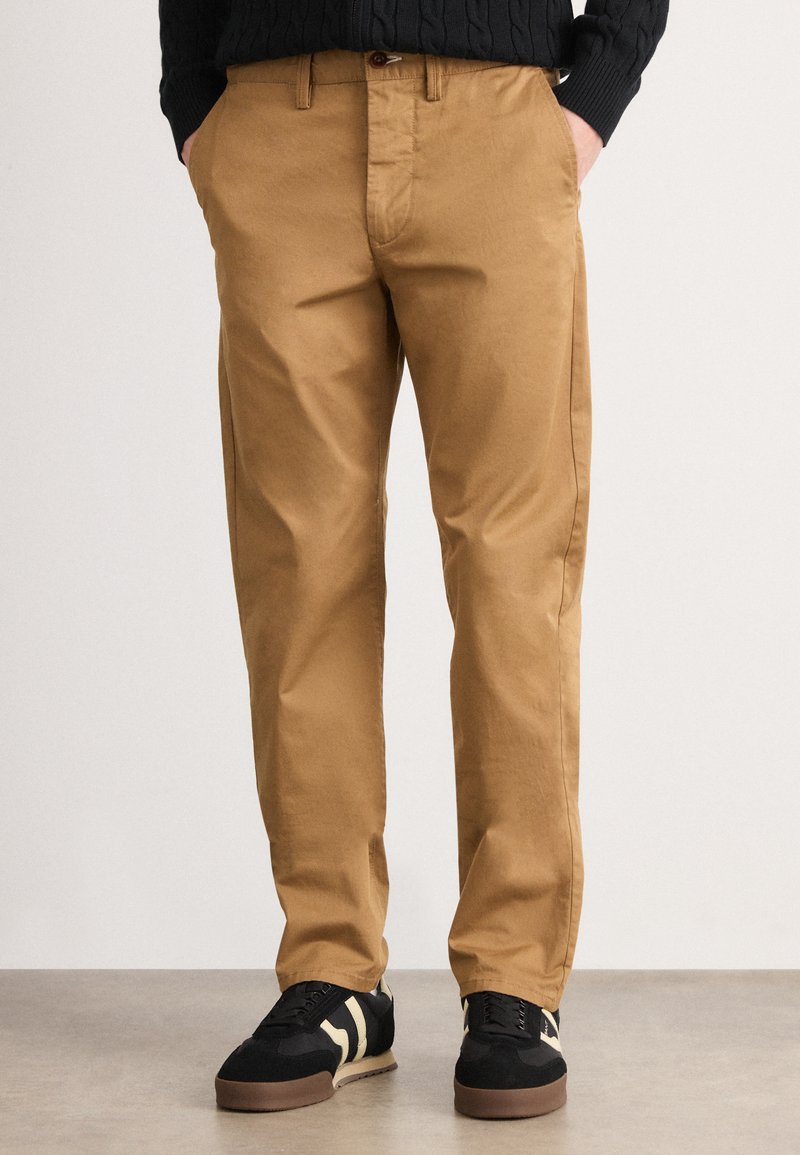 Mand iført lysebrune chinos med forlommer og sorte sneakers med beige striber, der står på et gråt gulv imod en ensfarvet baggrund.