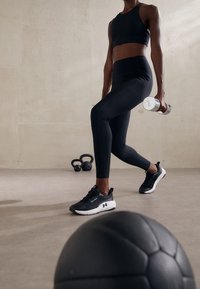 Černé sportovní oblečení zahrnující těsný crop top a legíny s vysokým pasem, doplněné černými běžeckými botami a stříbrnými činkami.