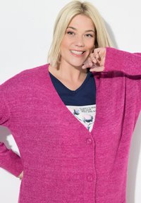 Cardigan rosa con una texture morbida, caratterizzato da scollo a V, maniche lunghe e due bottoni. Indossato sopra una camicia blu navy con grafiche floreali.