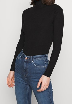 Personne portant un pull à col roulé noir moulant et un jean bleu taille haute, debout, les bras relâchés le long du corps.