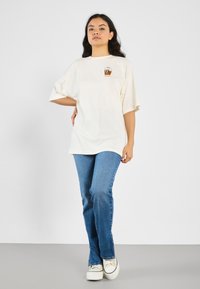 Krämfärgad oversized t-shirt med korta ärmar har en liten brun kopp-grafik. Bärs med blå utsvängda jeans och vita sneakers.
