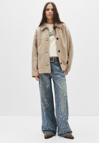 Beige suède jas met knopen, crèmekleurige top met logo, en wijdvallende blauwe jeans met vervaagde plekken en een lichte flared onderkant.