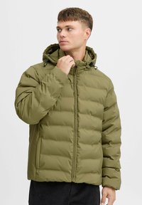 Olivenfarget puffjakke med hette, med kviltet design, frontglidelås og snøredetaljer. Lett materiale og komfortabel passform.