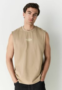Haut sans manches beige en tissu lisse avec un col rond et un logo minimaliste centré sur la poitrine. Silhouette ajustée.